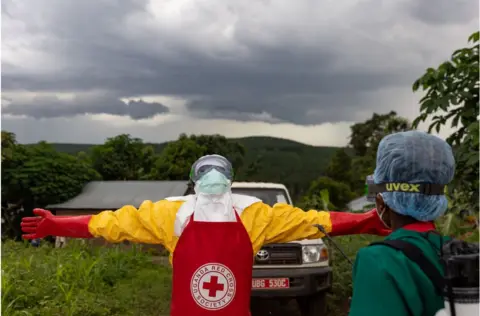 Getty Images Red Cross worker in personal protective clothes standing with their arms stretched out wide under very grey skies.
