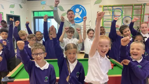 Children, mostly wearing blue school sweaters, and Kyren Wilson stand around a snooker table with their right arms up