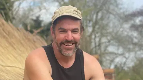 Nick Walker smiles at the camera while sitting on top of a thatched roof he had been working on. He wears a cream cap and a black tank top. A straw roof can be seen.