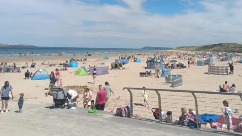BBC WEATHER WATCHER HELEN M Crowds of people on the beach with windbreakers, tents and towels on the sand. The sky is blue with white fluffy clouds and the sea can be seen in the background as waves crash against the shore.