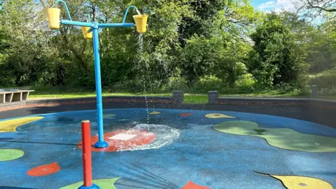 The splash pad in Ashbourne Park with water flowing out of yellow buckets on to a blue floor dotted with red, green and yellow areas