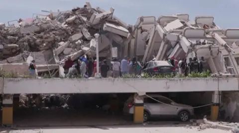 A collapsed building in Venezuela with people walking amid rubble and a car beneath them