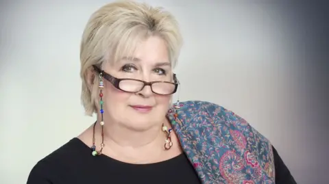 Dame Jenni Murray has a blue and red paisley pattened scarf over one shoulder and is wearing a black top, she has short hair and glasses which are perched on her nose and attached by a beaded string