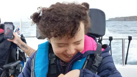 Family photograph Amelie, who has curly dark hair tied up in small bunches, is wearing a navy blue puffer coat and is smiling. She's on a boat with water visible behind her.