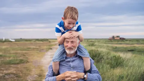 An man in his early 70s is wearing a blue shirt and a watch. He is carrying a young boy on his shoulders who is holding on to his head and smiling. There is a country road in the background and a blue evening sky.