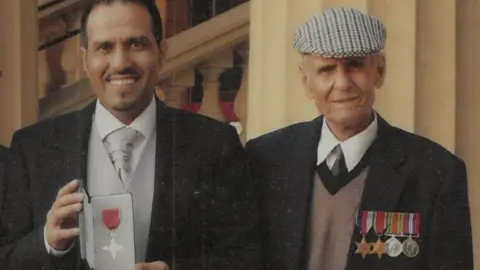 Mashuq Hussain Mashuq Hussain holding his OBE in grey and black tie and tails, standing next to  Ghulam Hussain wearing his medals  