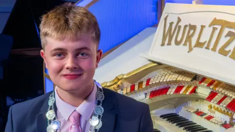 Damon Cox/Wurlitzer Heritage Trust Oscar, a 15-year-old standing next to an organ wearing a navy suit with a pink shirt and pink tie. He is looking directly at the camera and smiling.