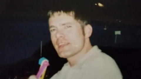 Supplied A grainy old image of Craig Rolfe, who has short brown hair and is holding a yellow brush or fan close to his face. He has his mouth partly open and is wearing a light shirt.
