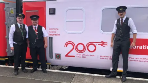 Three men in black and white uniforms with black caps standing beside a white railway carriage
