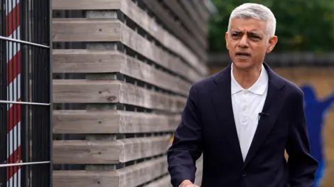 PA Media Sir Sadiq Khan walking down a London street. He is wearing a dark blue jacket and white shirt open at the collar. 