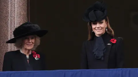PA Media Queen Camilla, wearing a broad-brimmed black hat with a black fur trim and a black jacket embellished with three red poppies, stands next to the Princess of Wales who wears a sculptured black hat, a black jacket and black lace scarf, with a silk red poppy on her lapel