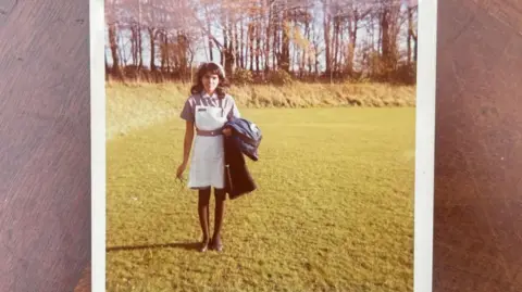 Savitri Cameron A photo of an old printed square photograph on a wooden table. The photo shows a young woman in a purple nurses dress and a white pinafore. She holds a pair of glasses in one hand and carries a coat in her other arm. She stands in an open grassy area where there are trees in the background