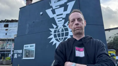 Cal is wearing a black sweatshirt with the words "Future Rebel". Future is in pink writing and rebel is in white writing. He has his arms folded and behind him is a grey building with a large design on it. 