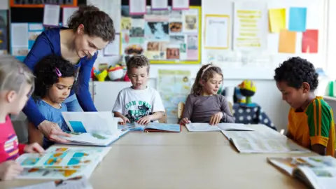 Getty Images Five children with text books sit around desks in a school classroom while a teacher helps one of the children with her book.