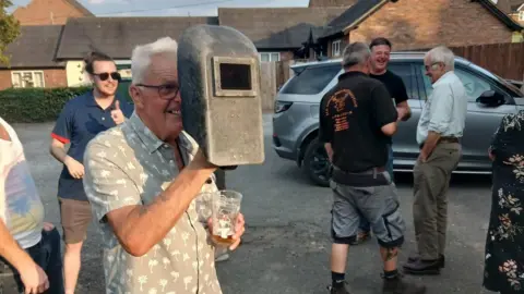 Kellie Wooldridge Glynne is wearing a brown shirt with a white palm tree pattern. He has grey hair and is holding a welding mask in front of his face with his right hand and has a pint glass in his left.