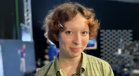 Alex Dunlop/BBC A head and shoulders image of a woman smiling at the camera. She has short auburn hair with two hair clips in, either side of her face. She is wearing a green checked shirt. 