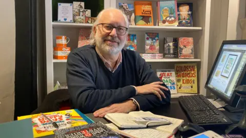 BBC / Naj Modak A bookshop owner sits at a desk, with books and a computer nearby. The man has a white beard and long white hair and is wearing black rimmed glasses. He has a pink shirt and a blue v-necked jumper on. 