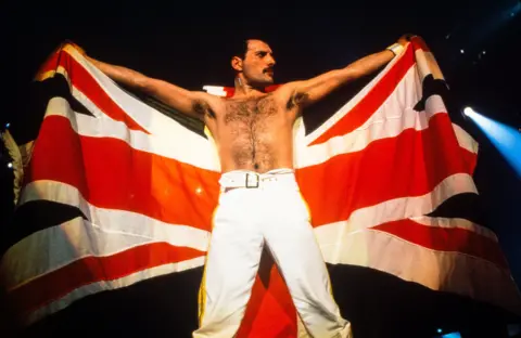 Getty Images Freddie Mercury, shirtless on the Knebworth stage, is holding a Union Jack behind him