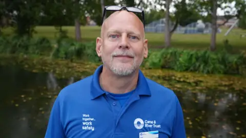 Dale Canfield has a bald head and glasses resting atop it. He is wearing a blue t-shirt and is standing in front of a canal.
