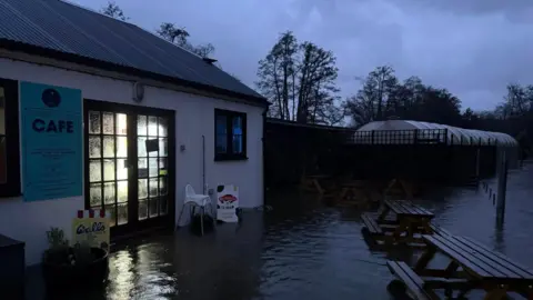A single storey building with a sign on it that says "cafe". The building, pictured in low light, is completely surrounded by water. Picnic benches, ice cream signs and a highchair are in the middle of the floodwater.