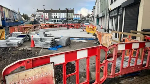 A construction site with a pile of tiles on the ground. It is surrounded by a red and white fence, along with cones.