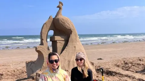Rachel Stubbs Cauley Yu stand side by side and smile in front of a sand sculpture of a peacock. The bird sits on top of a plinth, also made of sand. The sea can be seen in the background behind a long, sandy stretch of beach.