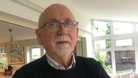 Tom Williams has a white goatee and glasses, and is wearing a shirt and blue jumper. The interior of a house with a glass beaded chandelier and window looking out to the garden behind can be seen in the background. 