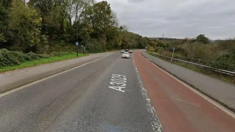 Google Maps A shot looking down an A-road, cars coming towards the camera. Three lanes, one for buses. Greenery on either side, a path and cycle path on one also.