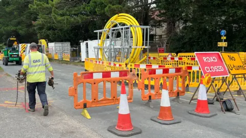 A workman in hi-vis clothing is carrying equipment next to bollards, plastic barriers, signs and a loop of yellow gas pipes