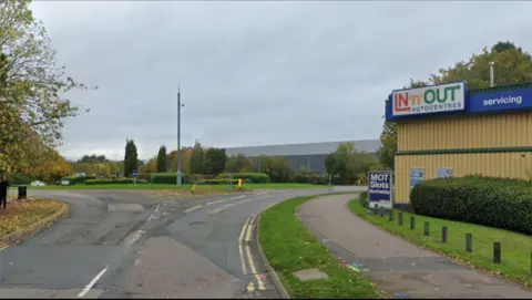 Industrial estate road leading onto a roundabout, which has trees and hedges in the centre. There is a large tree in the foreground, to the left, and a yellow warehouse building on the right which has a sign saying IN 'N' OUT AUTOCENTRES in multicoloured lettering.