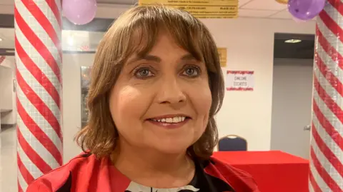 A woman standing in front of two red and white striped poles. She has short brown hair and is smiling. She's standing in a school corridor. There are balloons on the ceiling. 