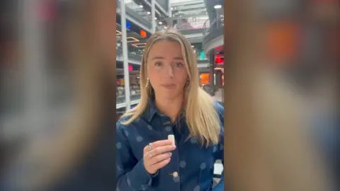 Lucy Woodham looks at the camera and holds a microphone with her right hand. She has long blonde hair and wears a denim dress with spots.