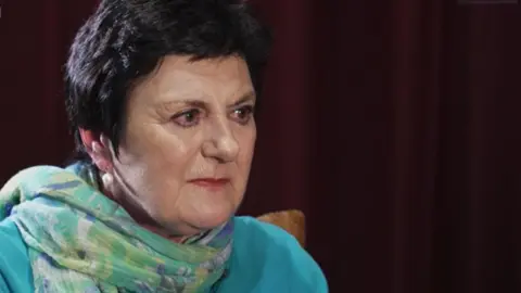 Ann Jensen sitting on a leather sofa in a dark room. She is wearing a blue cardigan and a turquoise scarf. She has dark hair in a short pixie cut, pearl earrings and dark pink lipstick on. She is looking at the interviewer to the right off camera.