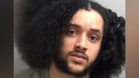 Essex Police A custody image of Ellis Heather, who has a lot of curly black hair. He is wearing a grey sweater.