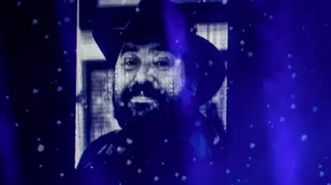 Picture of Gearóid Ó Cairealláin that was displayed on a screen in the SSE Arena during a gig by Kneecap - he has a dark beard and is wearing a wide-brimmed had