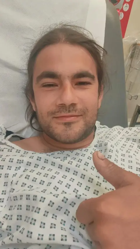 Sebastian Achim  in a hospital gown giving a thumbs up. 