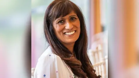 Lekha Desai Morrison A woman with long dark hair and wearing a white dress, looking sideways at the camera and smiling