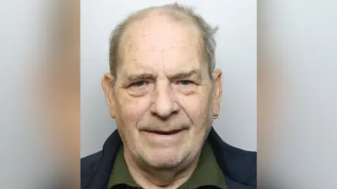 A mugshot of Sunter who appears to smile at the camera with no teeth. He has a weathered face, wispy grey hair and white facial stubble. He is wearing an earring in his left ear and is dressed in a dark green top and navy coat or blazer.