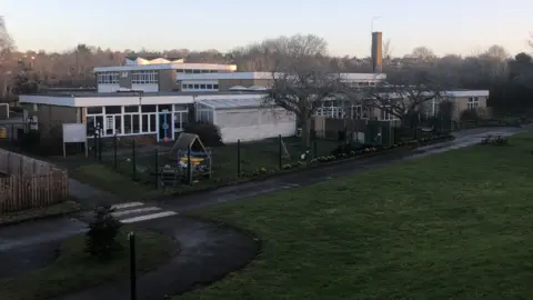 St John's Catholic Primary School A wide view of a low-rise school building surrounded by grass, trees and paved paths on a clear day. Several connected blocks with flat roofs are visible, along with fenced play areas and outdoor equipment.