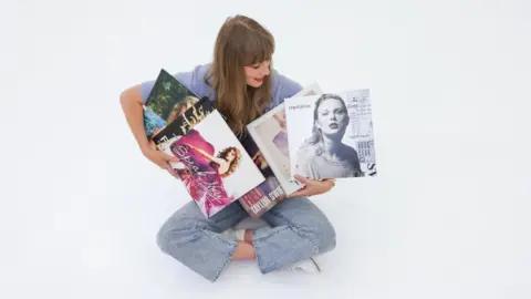 Taylor Swift Taylor Swift sits cross-legged on the floor, holding copies of her first six albums