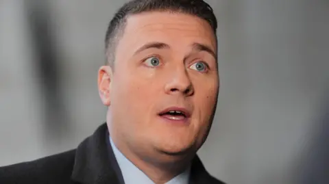 PA Media Health Secretary Wes Streeting with short brown hair and blue eyes wearing a suit and black coat.