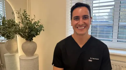 Tom is sat in his clinic, smiling at the camera. He is wearing a black, scrubs-style top, with his name embroidered on the left. To the left of the picture is a plant pot on a pedestal, and a mirror. Behind Tom is a window. It is a head and shoulders shot of him.
