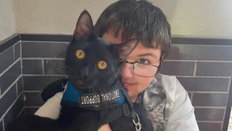 Family handout A boy with short dark hair and black rimmed glasses sat on a chair cuddling a black cat, which has a harness around it with the words 'emotional support' on it. The boy is wearing a grey T-shirt and sat against a wall inside.