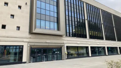 BBC Exterior view of the newly redeveloped former Debenhams building in Gloucester, with a light modern façade, large windows and paved pedestrian area in front.

