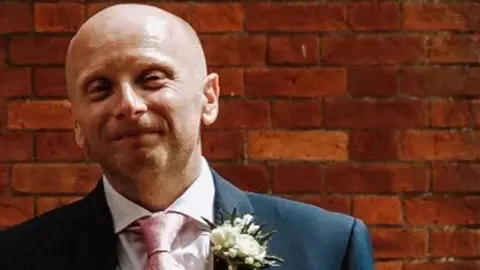 Dan Kay smiling wearing a suit with pink tie and white flowers on the lapel