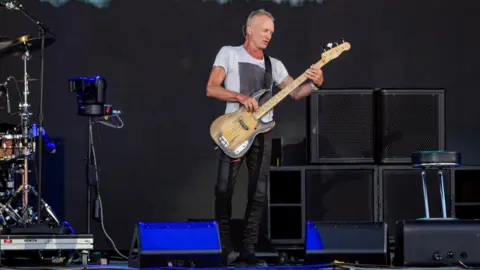 PA Sting performing at the Isle of Wight Festival on Friday. He has short, cropped grey hair and wears a white t shirt and tight dark trousers. He's playing a bass guitar.