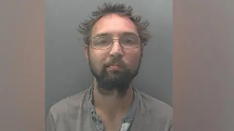 Cambridgeshire Police Police mugshot of a man with grey sticky-up hair and glasses. He is wearing a grey open-necked shirt