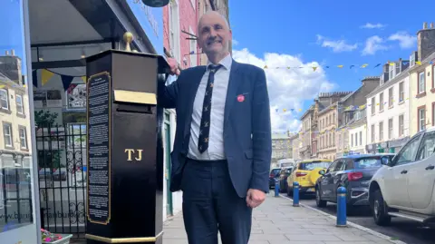 A bald-headed man wearing a suit leans against a black post box, with a row of cars behind him