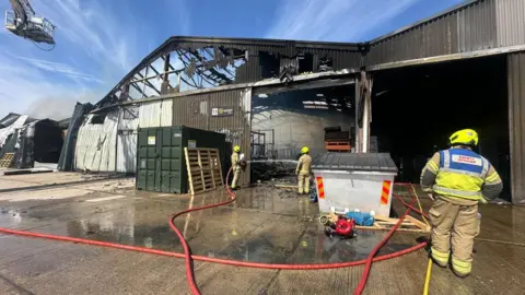 Firefighters in yellow outfits with long red hoses damping down at a warehouse with its roof blackened and exposed with fire damage