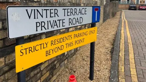 Robbie Kalus/BBC A white rectangular sign saying "Vinter Terrace Private Road" in black capital letters. Below is a yellow rectangular sign saying "strictly residents parking only beyond this point" in black capital letters. The signs are fixed to two black posts which stand in brown gravel next to a paved road marked with double yellow lines. 